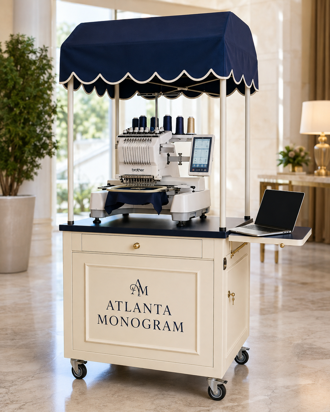 A cream-colored mobile embroidery cart with a navy blue scalloped canopy, featuring an industrial embroidery machine, a side laptop tray, and the text 'Atlanta Monogram' on the front, set in a bright, modern interior.