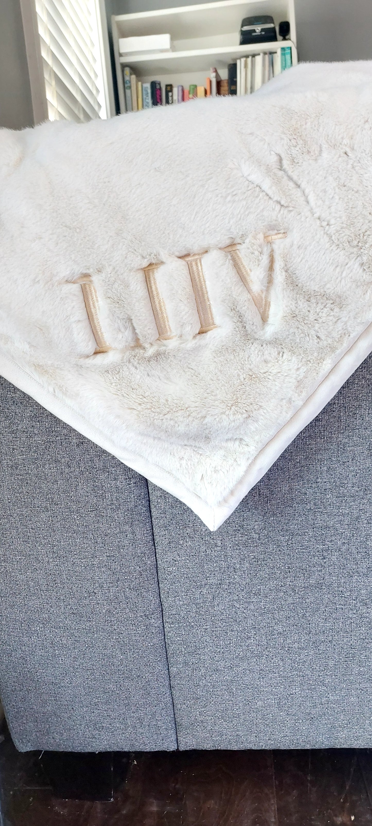 A custom embroidered blanket with the initial 'L' on it, draped over the arm of a gray sofa, with a part of the blanket folded to reveal the embroidery.