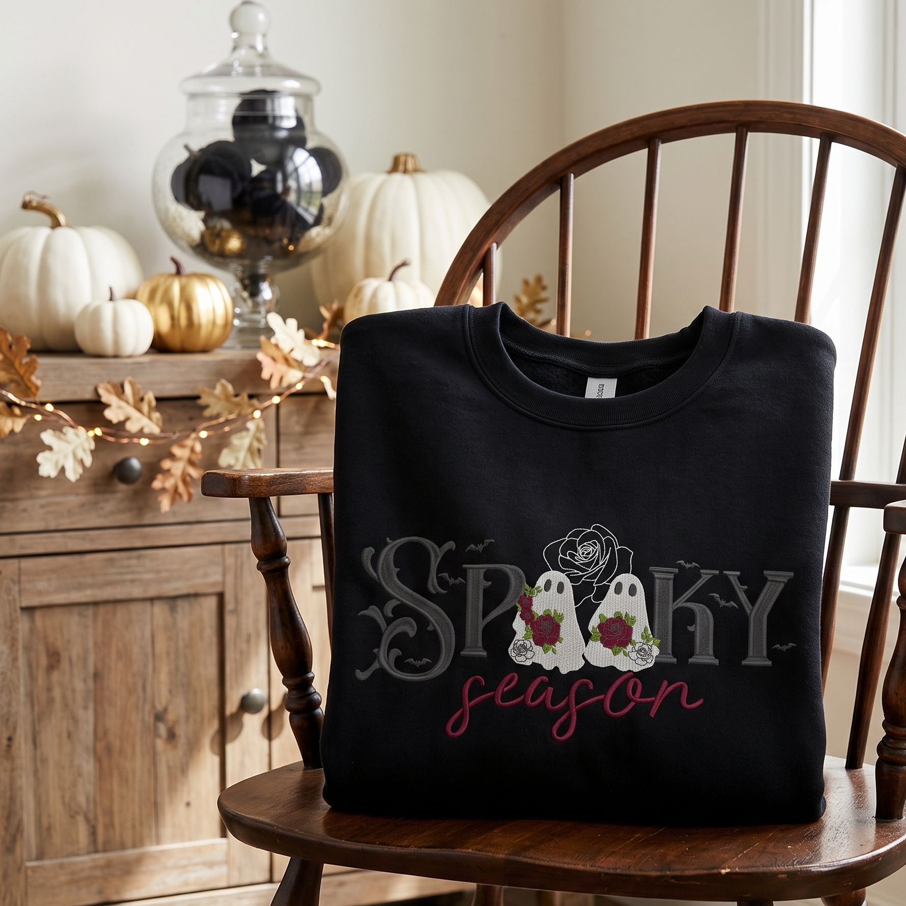 A black crewneck sweatshirt with a 'Spooky Season' graphic sits folded on a wooden spindle-back chair in a cozy, autumn-themed room with pumpkins and a window.