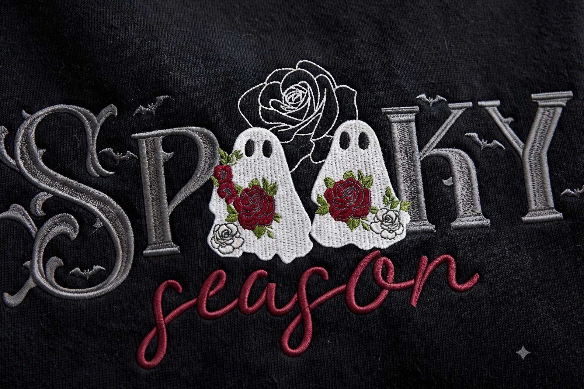 Close-up of embroidered text reading 'Spooky season' on a black fabric background, with two white ghost figures decorated with red and white roses.